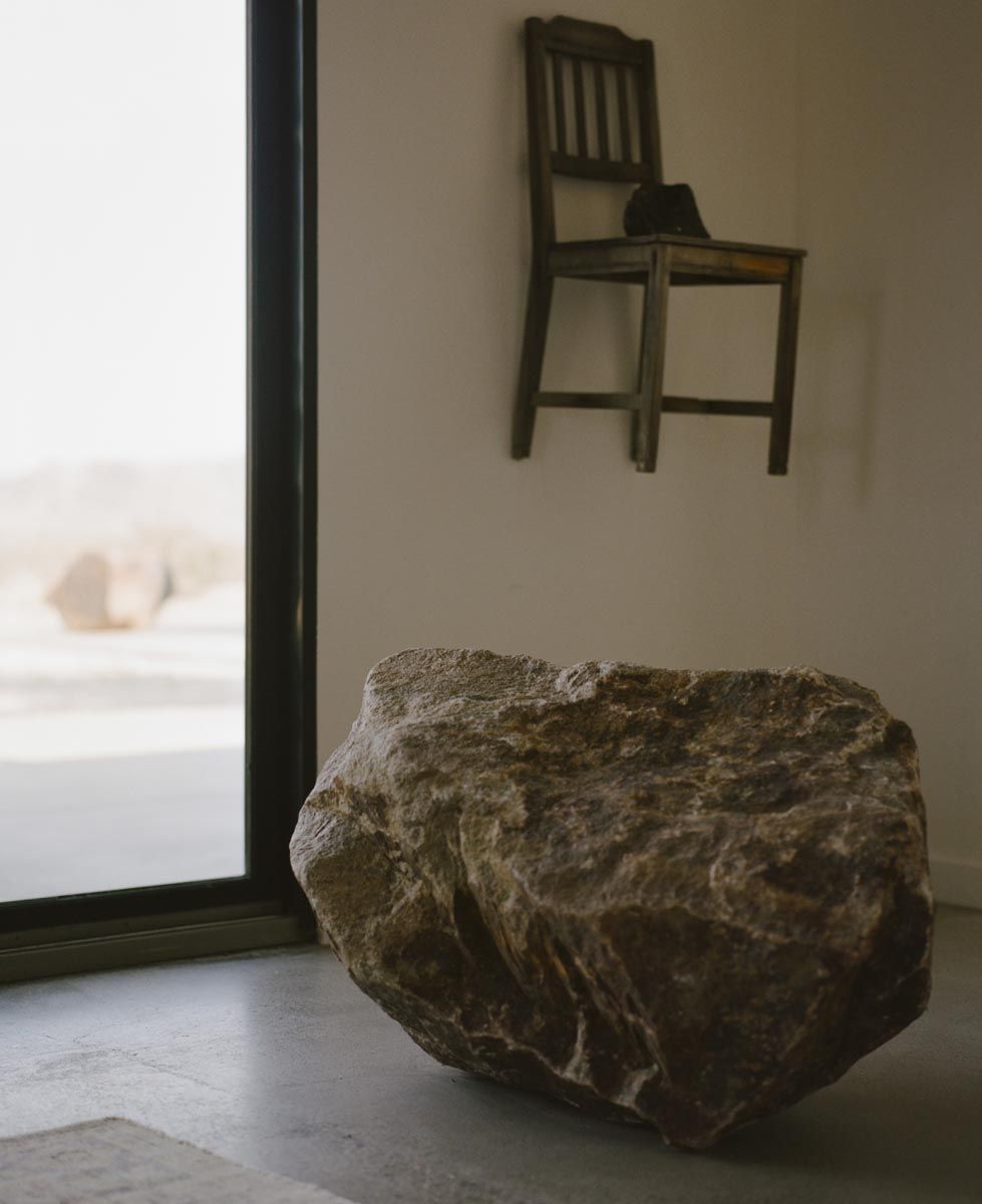 A boulder resting on the floor by a window, a chair hung on the wall above, at Goat Mountain House, Joshua Tree