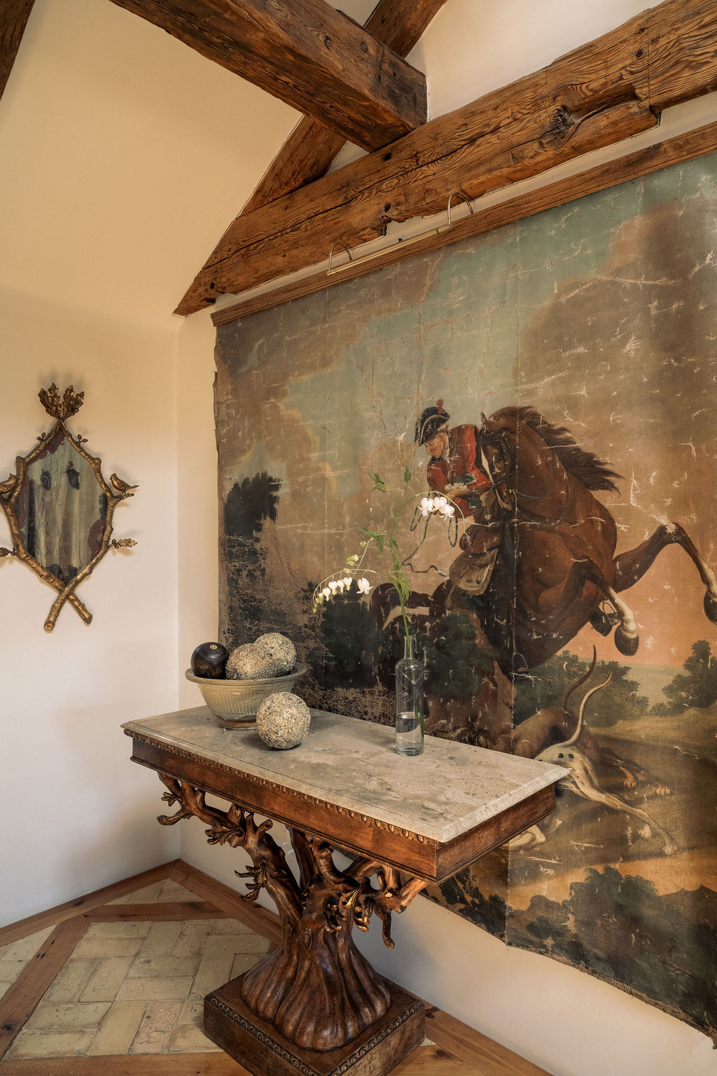 Antique hunt-scene canvas above a carved tree-trunk console under hand-hewn timber beams