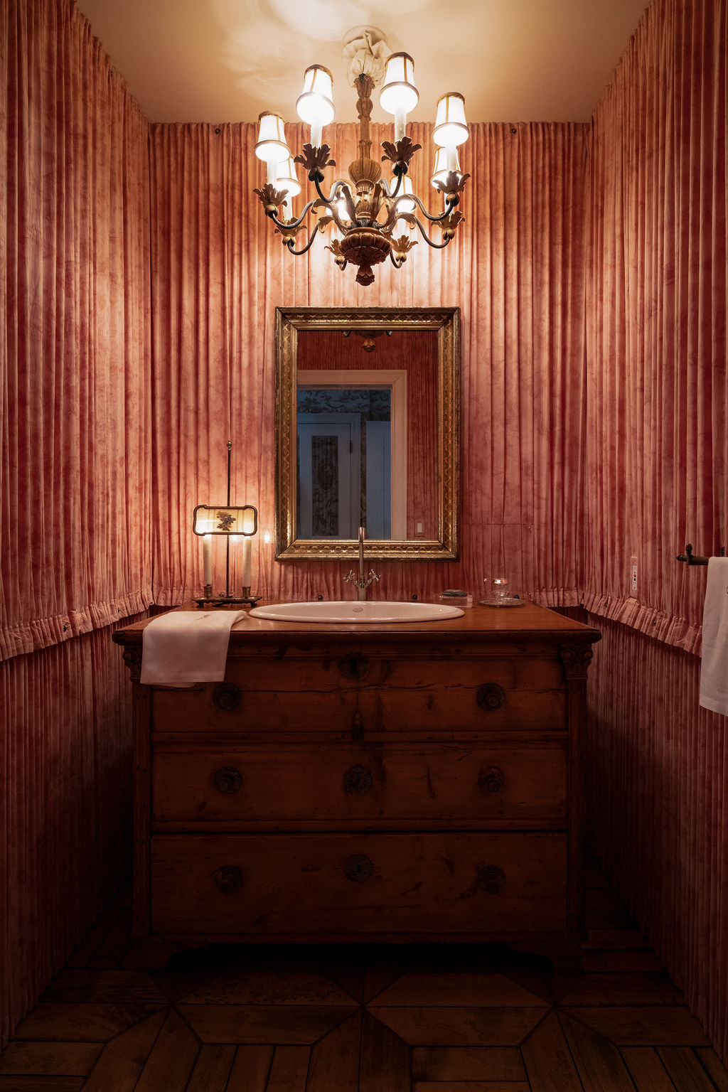 Powder room lined in pleated rose fabric with a gilt chandelier and an antique commode sink